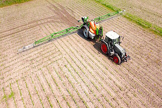 Grauer Traktor mit grünem Spritzgestänge fährt auf neu bepflanztem Feld.