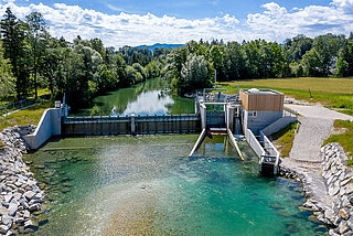 Pequeña central hidroeléctrica con esclusa y cabina de mando, representada en forma de diagrama con cielo azul y pequeña área verde. 
